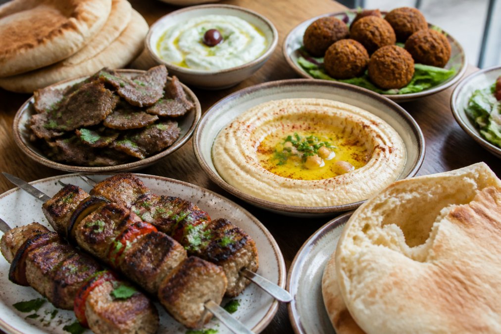 A spread of authentic Lebanese and Mediterranean dishes arranged on a rustic wooden table: a ceramic bowl of hummus drizzled with olive oil, crispy golden falafel, thinly sliced gyro meat, a small dish of tzatziki, grilled kabobs with char marks, and warm pita bread torn open to show the soft interior, natural light from a nearby window