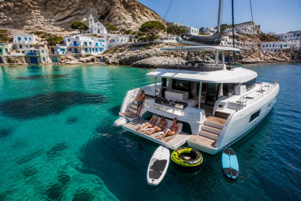 Sailing catamaran anchored in a turquoise cove surrounded by white-washed buildings and rocky cliffs of a Greek island, guests relaxing on the yacht's teak deck with water toys visible, clear Mediterranean waters and blue sky