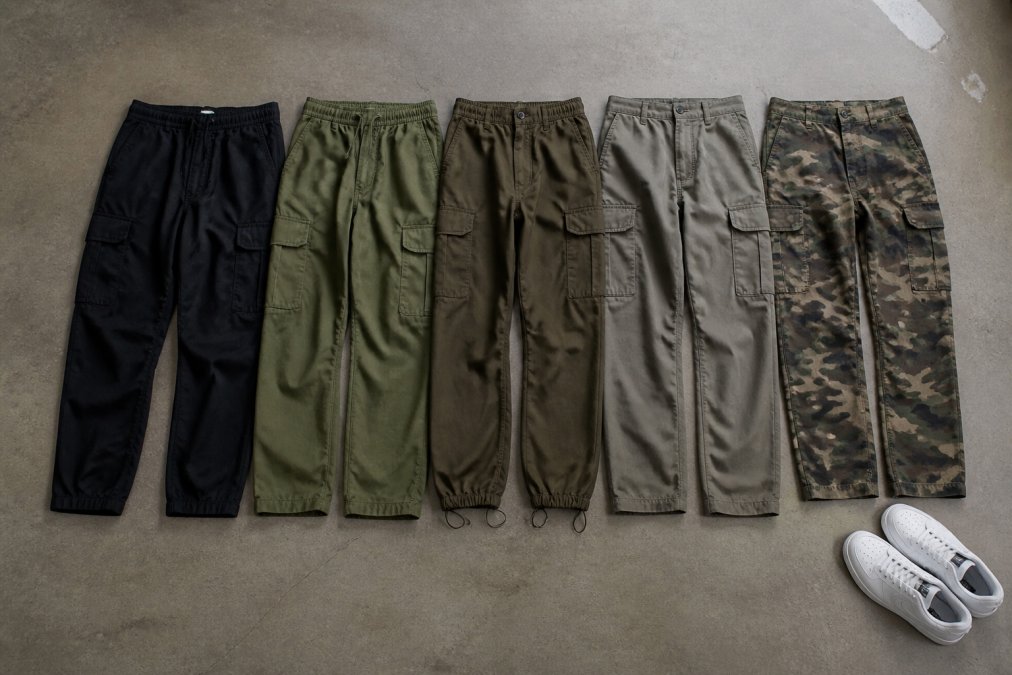 A flat lay of five different pairs of men's cargo pants in black, light olive, and olive camo patterns, arranged neatly on a polished concrete floor with a pair of white sneakers at the edge