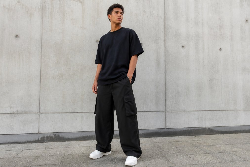 A stylish young man wearing an oversized black t-shirt and wide-leg cargo pants, standing against a concrete urban wall, looking relaxed and confident in natural daylight