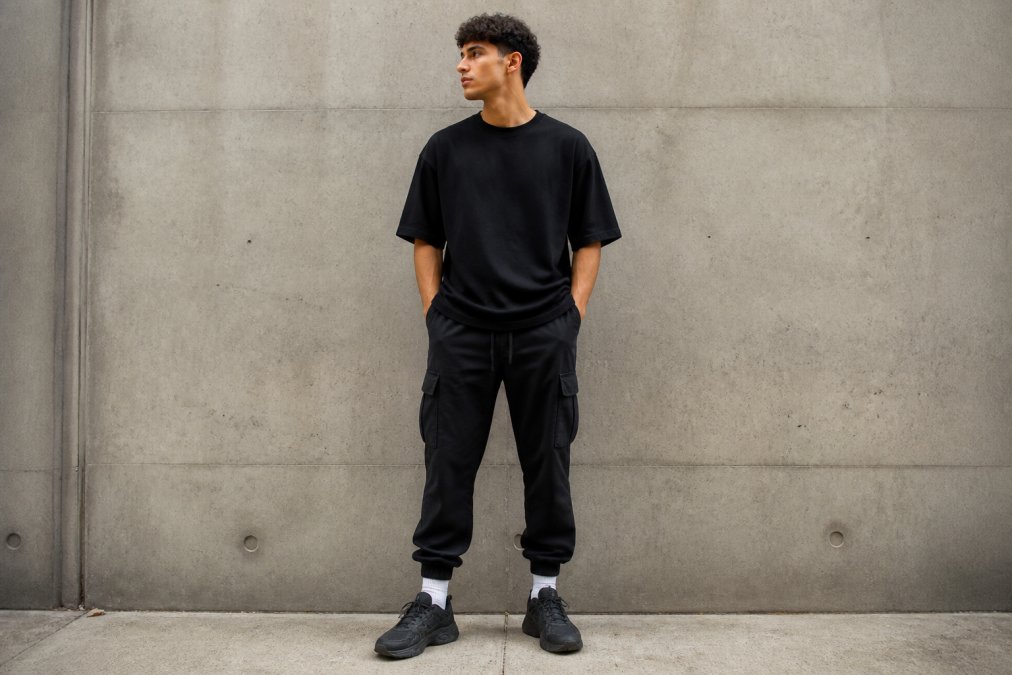 Young man in a black oversized streetwear t-shirt and cargo joggers posing against a concrete wall.