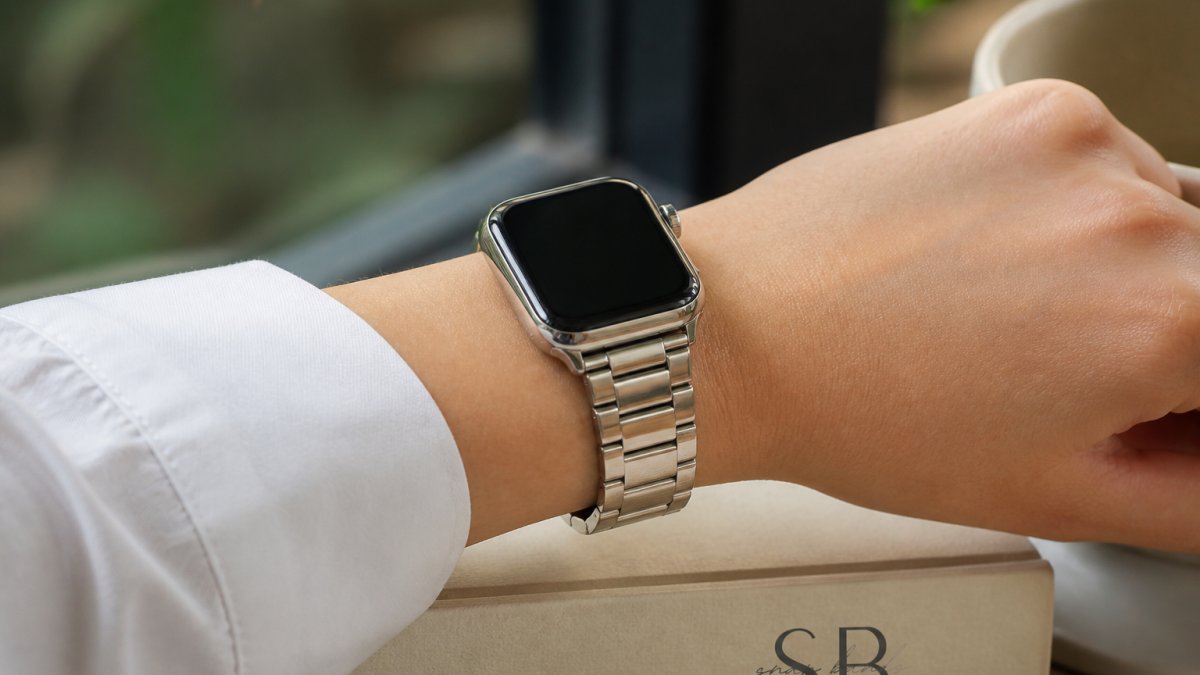 Woman's wrist wearing a polished stainless steel metal band on an Apple Watch, photographed in natural daylight showing the band's refined finish and secure fit against her skin
