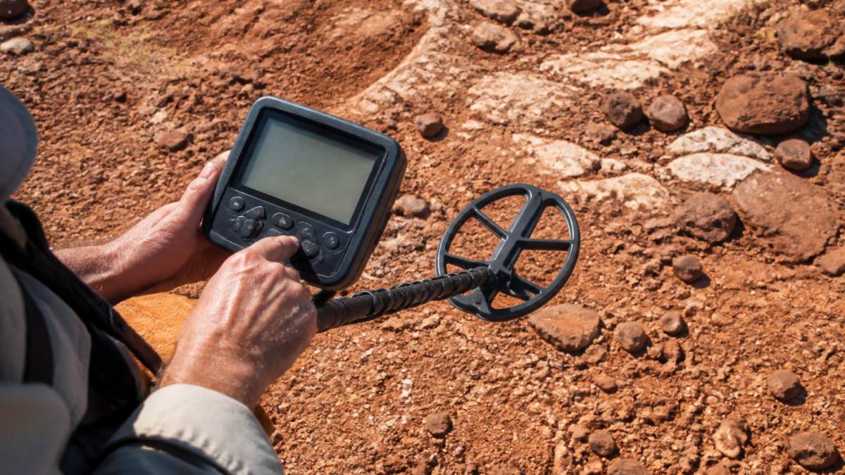 Close-up of prospector's hands adjusting ground balance controls on a pulse induction detector display screen in the field, with red mineralized soil and exposed bedrock visible in the background