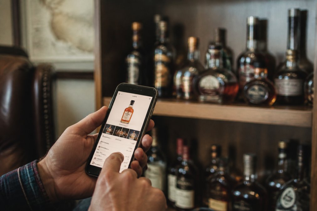 Person holding smartphone displaying whiskey collection app with bottle details, tasting notes, and inventory list visible on screen in home setting