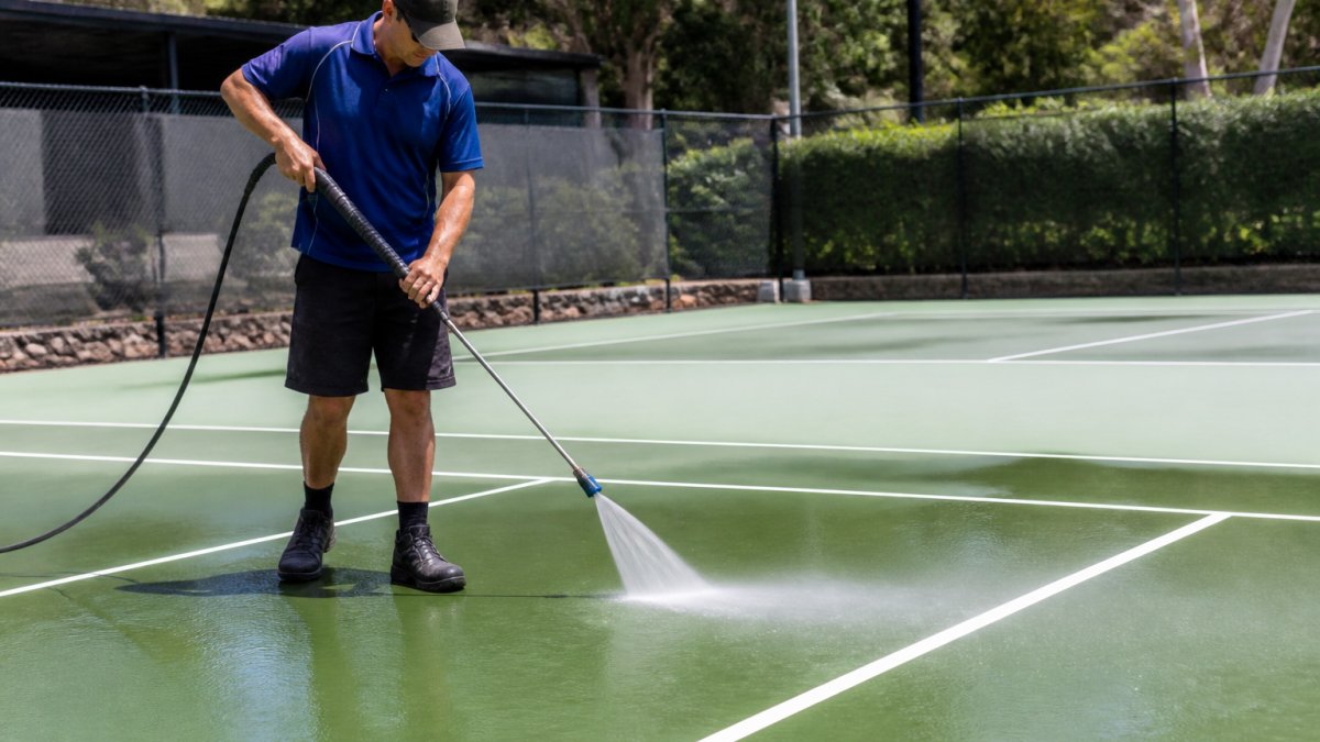 A professional cleaner using a low-pressure soft wash wand on a green acrylic tennis court surface, with visible water spray and clean line markings in the foreground
