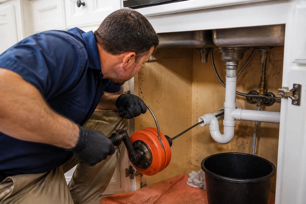 Professional plumber's hands operating a manual drain snake with proper grip on the crank handle, inserting the flexible steel cable into a kitchen sink P-trap with focused concentration