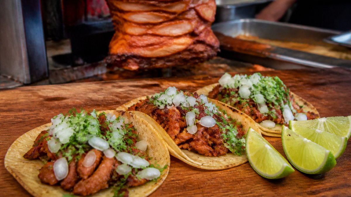 Close-up of freshly prepared tacos with al pastor meat, diced cilantro, white onions, and fresh lime wedges on a wooden counter at a taqueria