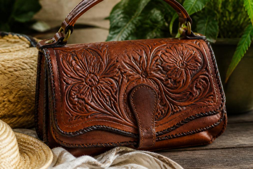 Close-up of a richly textured, buttery-soft full-grain leather handbag with hand-tooled floral detailing resting on a rustic wooden surface, warm natural light highlighting the supple texture and hand-stitching