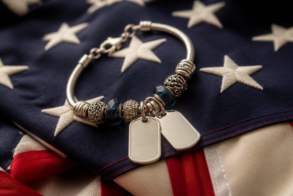 Close-up of a sterling silver charm bracelet with military dog tag charms and decorative beads laid on a folded American flag, warm soft lighting