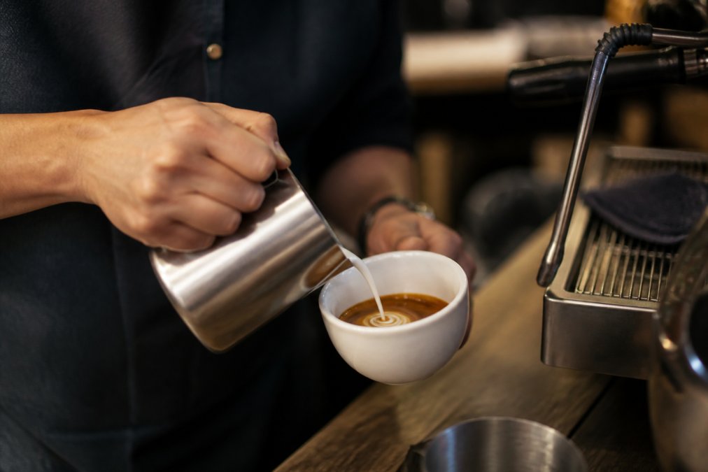 Close-up of a barista's hands holding a stainless steel milk pitcher, pouring microfoam into a white ceramic cup of dark espresso on a wooden counter, steam wand visible in the background