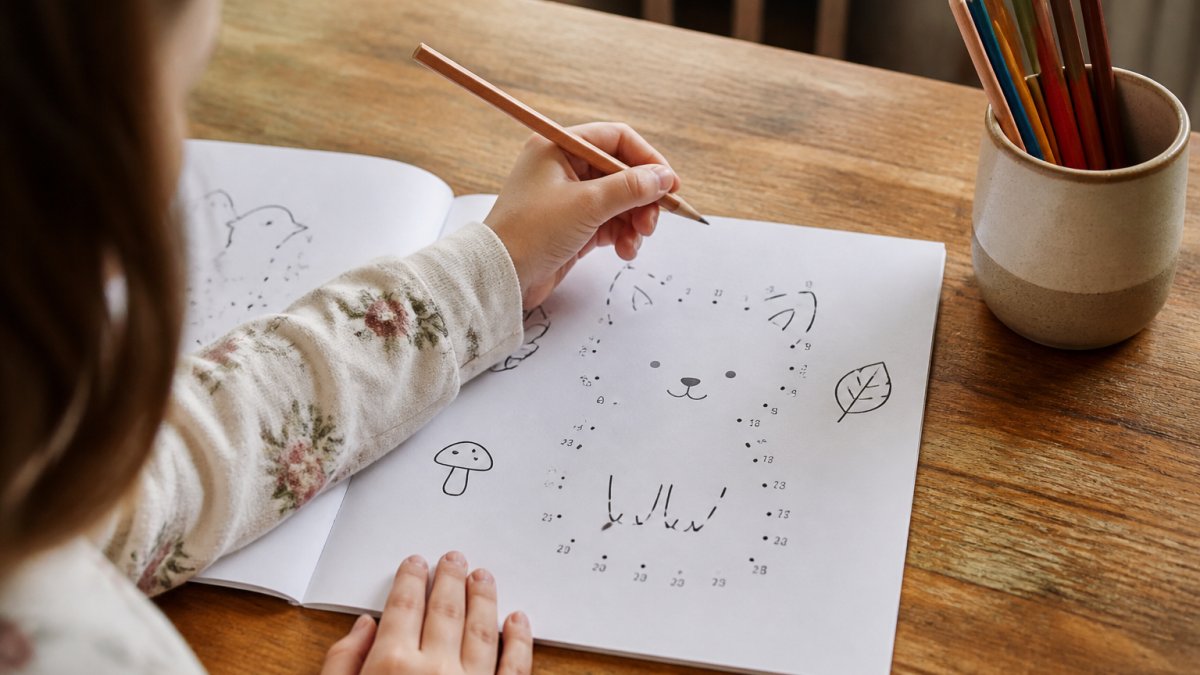 Child's hands holding a pencil over a minimalist dot-to-dot activity page with simple line drawings, sitting at a wooden table with colored pencils in a cup nearby