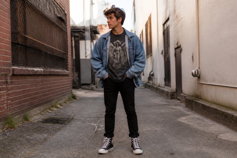 Man wearing a minimalist graphic tee layered under an oversized denim jacket, styled with tapered black jeans and high-top sneakers, standing in an urban alleyway with natural morning light