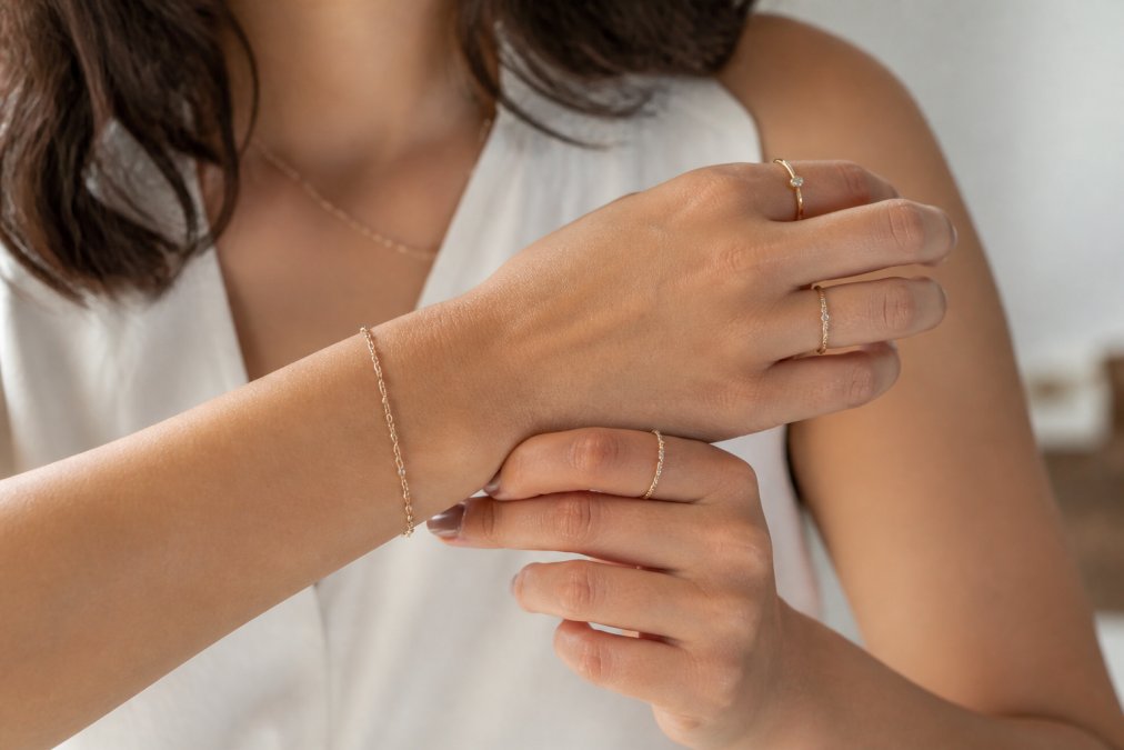 Close-up of a woman's hands and wrist wearing layered minimalist jewellery, three delicate stackable rings on different fingers and a fine-link chain necklace visible against her wrist, demonstrating everyday elegant styling in natural daylight