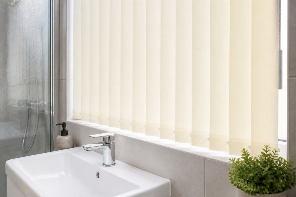 Close-up of white PVC vertical blinds installed above a modern bathroom sink, showing the wipe-clean fabric texture and flexible slats in a bright, steamy bathroom environment