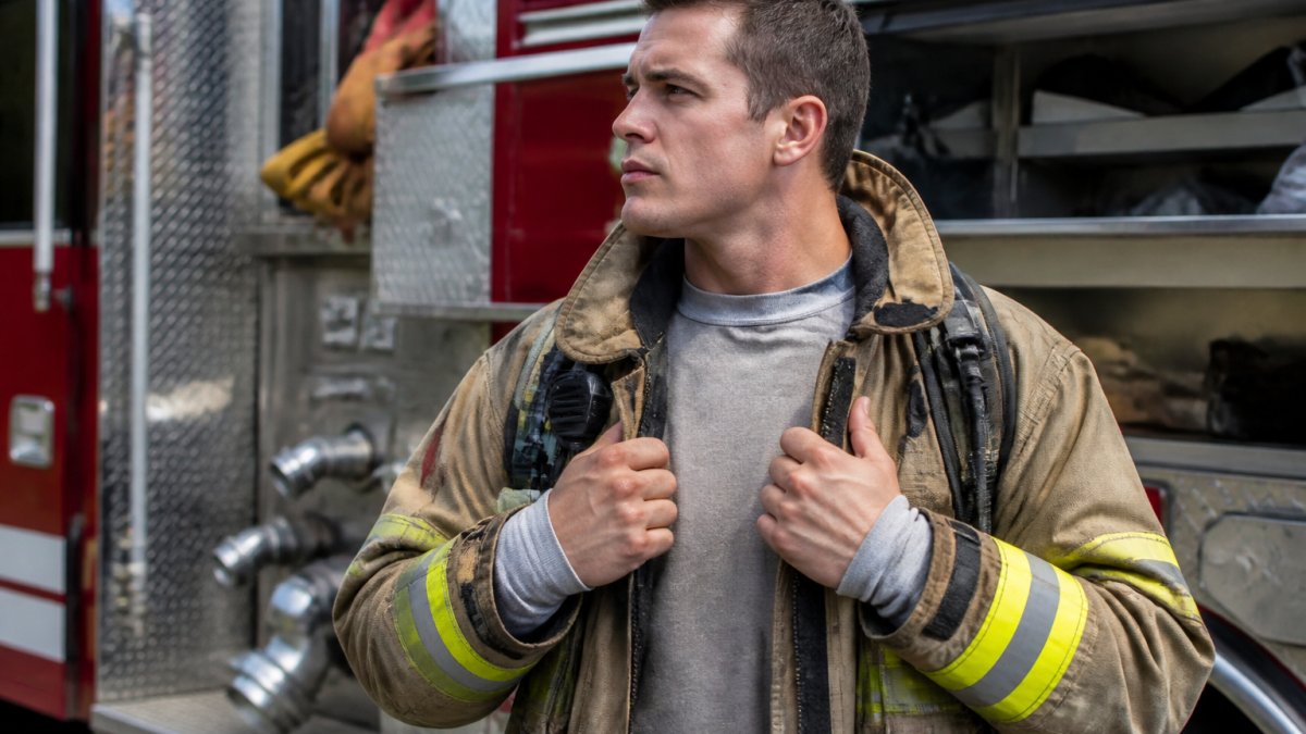 Firefighter in full turnout gear standing in front of fire engine, showing visible base layer at neck and wrists with moisture-wicking material