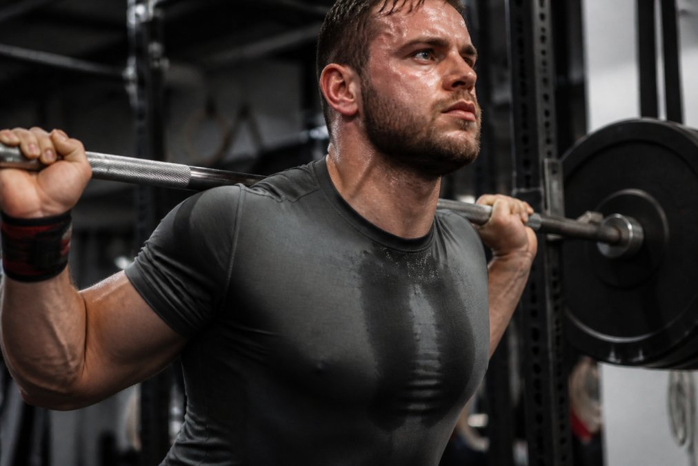 Close-up of a lifter mid-heavy squat wearing a moisture-wicking performance shirt, sweat visible on the fabric showing effective moisture transport, muscular physique visible through athletic fit