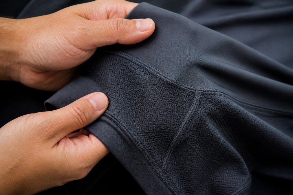 Close-up of performance fabric showing flatlock seams and breathable mesh panels with hands touching the material to demonstrate texture and construction