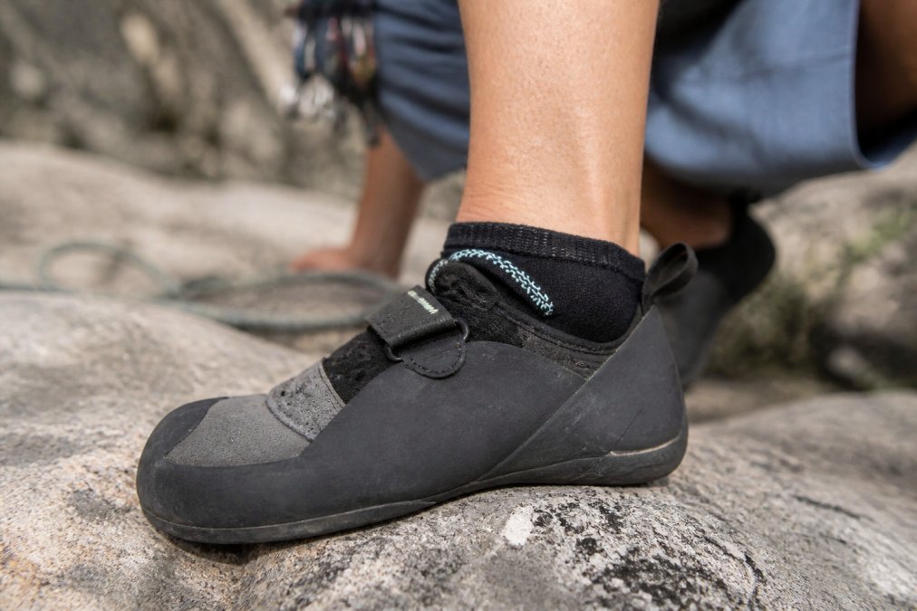 Close-up of a climber's foot in a tight climbing shoe with a moisture-wicking sock visible at the ankle, showing proper fit and sock positioning during a rest break on granite