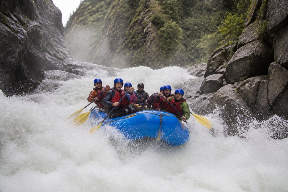 A raft packed with six paddlers in helmets and PFDs plunging through a churning whitewater rapid in spring, surrounded by steep green canyon walls with mist rising from the water