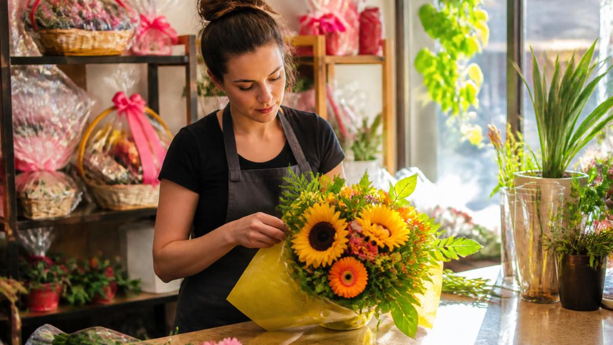 A florist in a bright shop arranging a vibrant birthday bouquet of sunflowers and gerbera daisies, wrapped gift baskets visible on a shelf behind the counter, warm afternoon light through the window