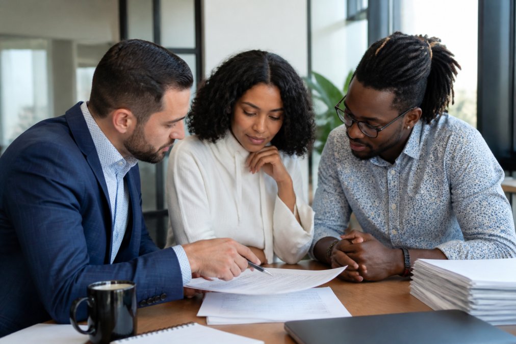 Professional loan officer and diverse couple reviewing mortgage documents together at modern office desk with natural lighting