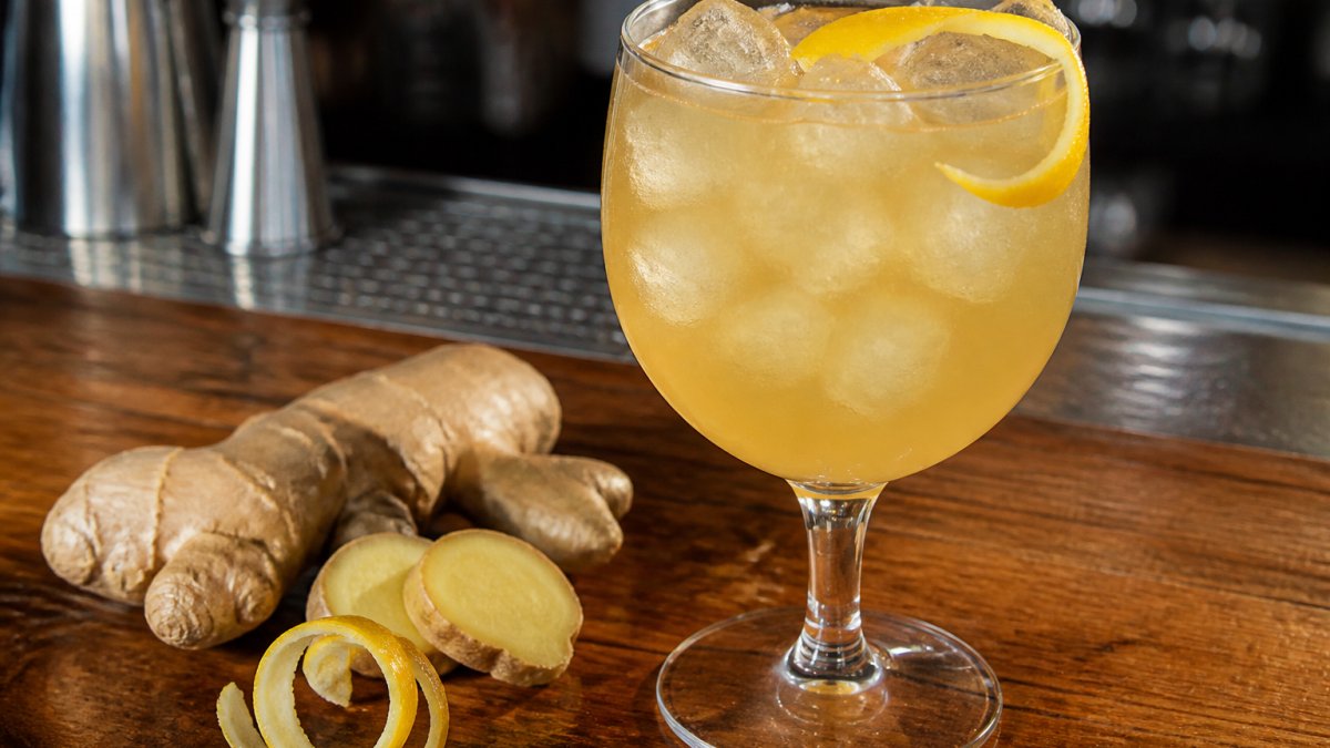Close-up of a chilled non-alcoholic ginger aperitif poured over ice in a stemmed glass, with a fresh ginger root and lemon twist beside it on a wooden bar top