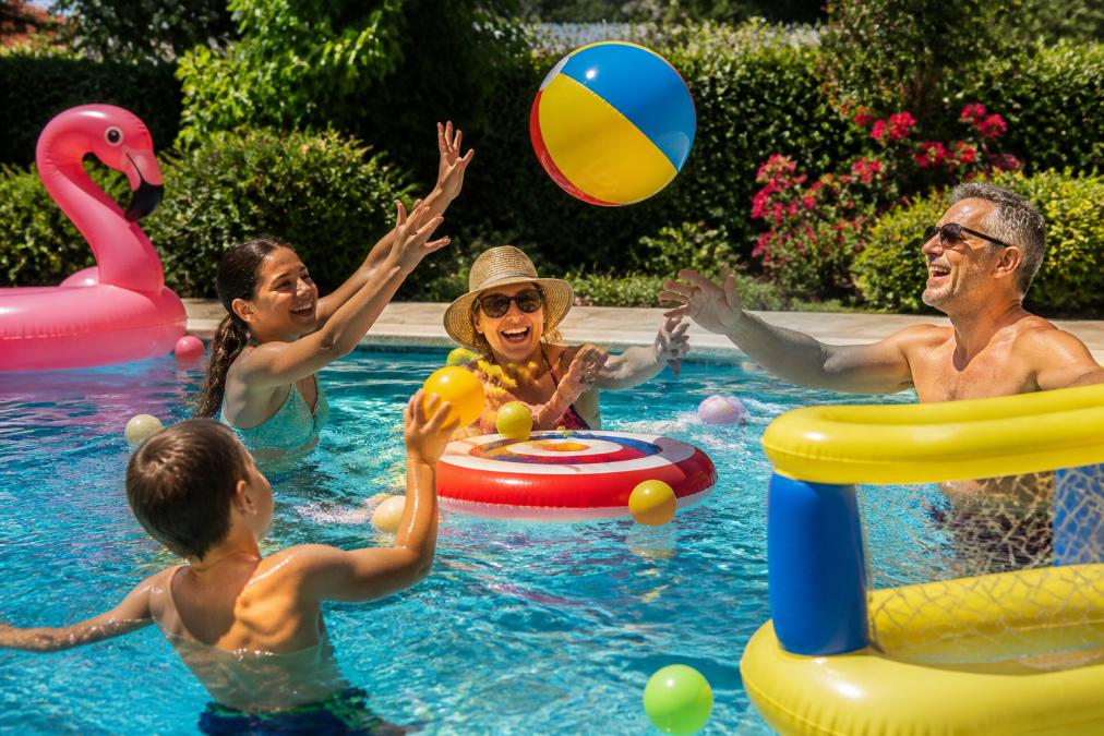 Multigenerational family laughing and playing with colorful inflatable pool games together in a sunny backyard swimming pool, with children and adults tossing balls and reaching for floating toys