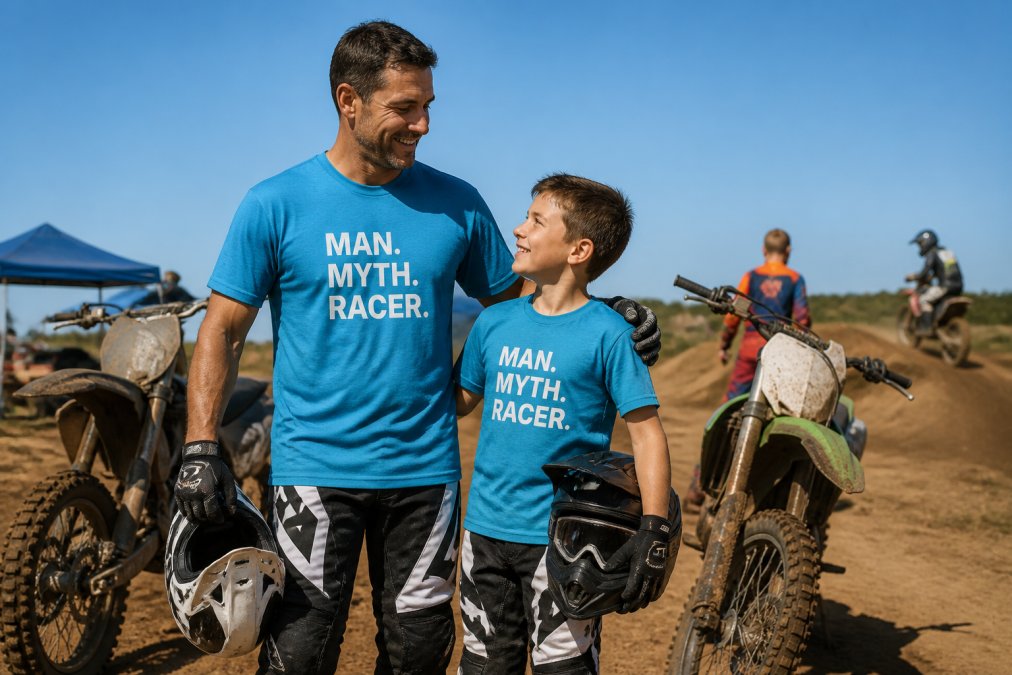 Father and son at a motocross track day event standing next to their dirt bikes, both wearing matching motocross gear, with the track and other riders visible in the background under bright daylight