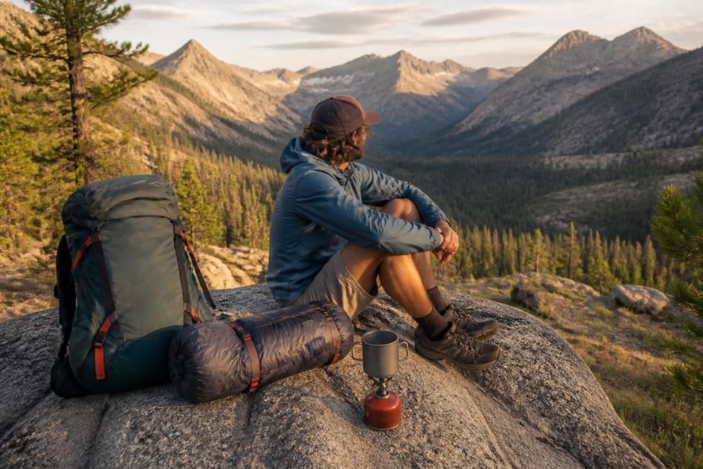 A hiker in their 30s sitting on a granite boulder at a mountain campsite, surrounded by a fully packed backpack, rolled sleeping quilt, and a small stove, with a valley and distant peaks visible behind them in warm late-afternoon light