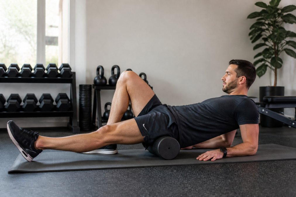 Athlete in athletic wear demonstrating foam roller technique on hip flexor area while lying on the floor in a home gym setting with natural lighting from windows