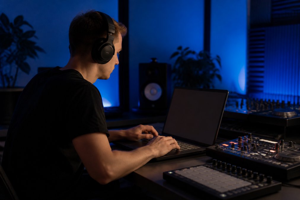 Music producer working at laptop with headphones and mixing equipment in home studio, reviewing distribution analytics on screen with blue lighting