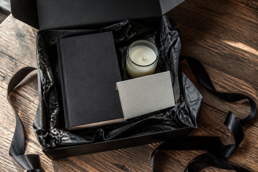 A curated gift box open on a wooden table, containing a dark-spined hardcover novel, a small glass candle, and a folded cream note card, surrounded by dark tissue paper and a black satin ribbon, photographed from directly above in natural window light