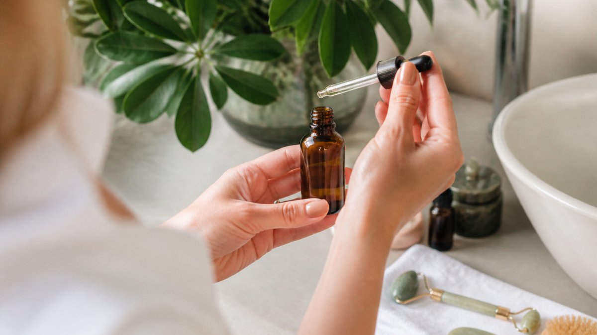 Close-up of a woman's hands holding a small amber glass serum bottle with a dropper, soft natural light, green botanical leaves blurred in the background, clean minimal bathroom counter
