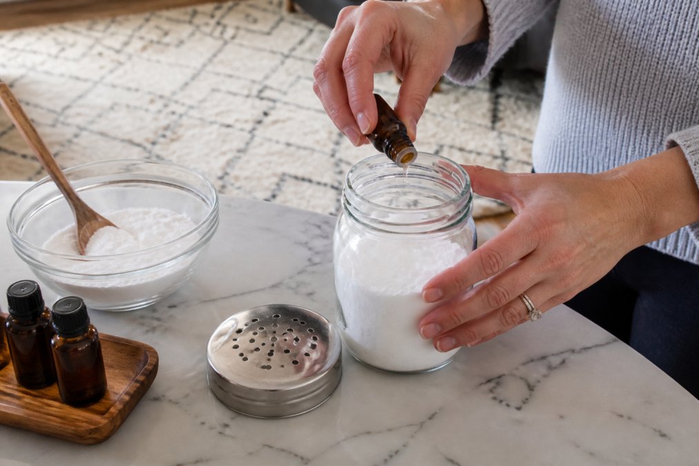 Close-up of hands mixing baking soda and essential oils in a glass jar on a kitchen counter, with a shaker lid nearby and a carpet in the background