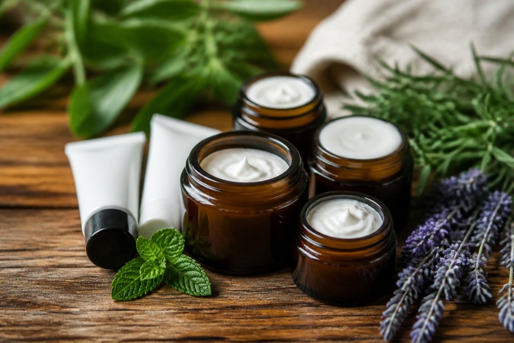 Close-up of several small amber glass jars and cream tubes arranged on a wooden surface with green leaves and lavender sprigs nearby, soft natural lighting