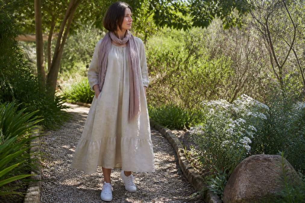 Woman wearing a natural linen peasant dress in neutral tone, styled casually with white sneakers and a linen scarf, standing in soft natural daylight in a garden setting