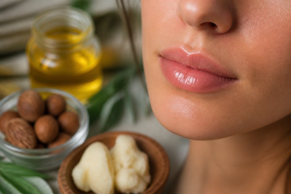 Close-up of woman's lips with radiant natural gloss applied, showing luminous shine and hydration, with botanical ingredients like jojoba oil and shea butter visible in soft focus background
