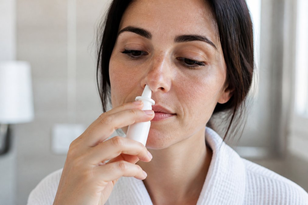 Close-up of hands holding a nasal gel bottle with applicator tip positioned at the entrance of a nostril, demonstrating proper angled insertion technique and gentle application in a bright, clinical setting