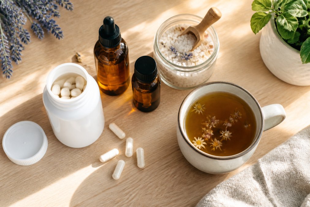 Overhead flat lay of natural relaxation products on a light wood table: a bottle of magnesium capsules, amber essential oil bottles, a small jar of bath salt, and a cup of herbal tea, with soft natural window light