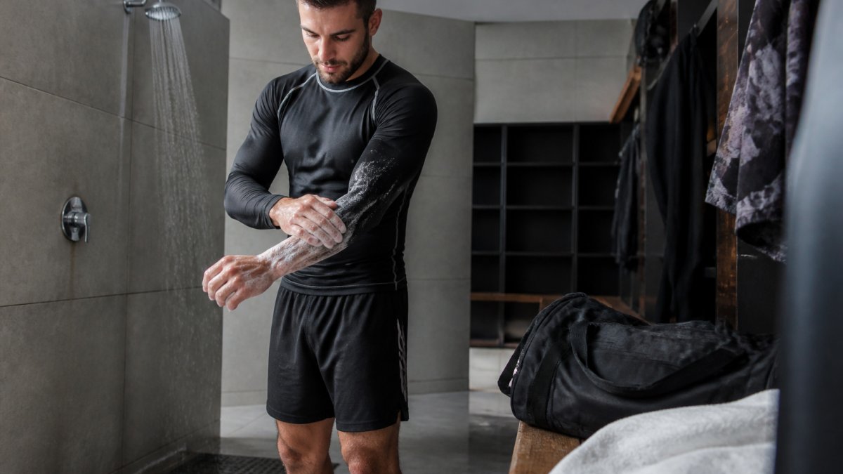 A grappler in a rashguard and shorts standing in a locker room shower area, washing hands and arms with soap, with a gym bag and clean towel nearby