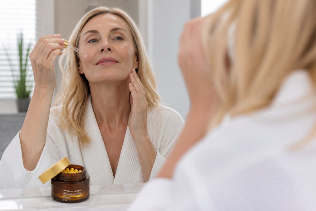 Woman in her 50s applying natural skincare products in a bright, minimalist bathroom with soft morning light, showing her applying serum to her face and neck area in the mirror