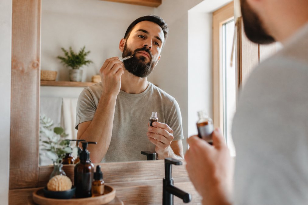 Man with a groomed beard applying natural skincare oil in a bright, modern bathroom mirror.