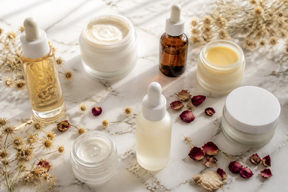 Flat-lay of several glass and minimal-packaging skincare bottles and jars arranged on a white marble surface with dried chamomile flowers, rose petals, and soft natural window light