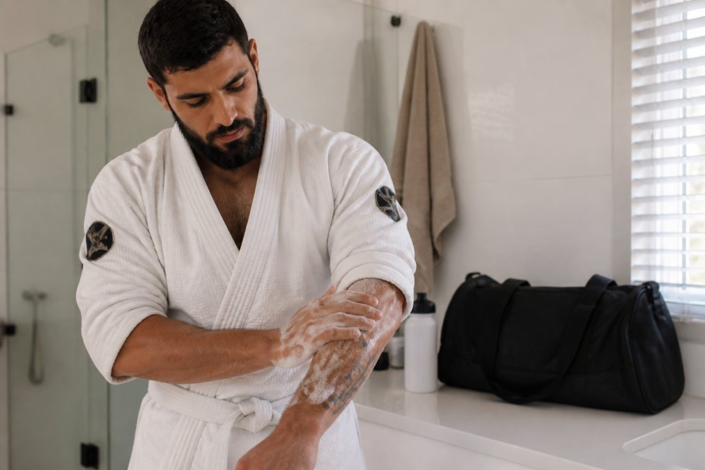 A martial artist in a white gi standing in a clean, bright bathroom, applying soap lather to their forearm after training, with a gym bag and water bottle visible on the counter behind them