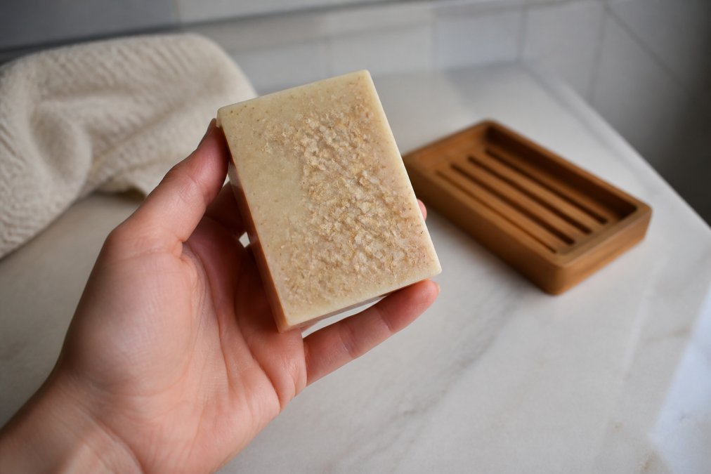 A hand holding a natural beige soap bar with visible oatmeal flakes, soft bathroom lighting, wooden soap dish in background