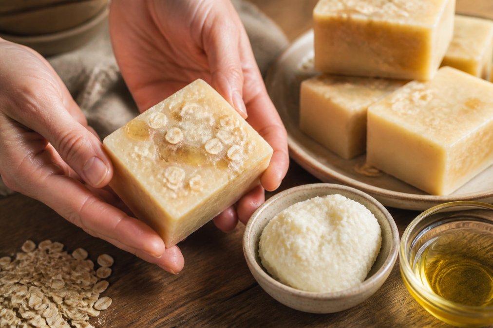 Hands holding a handcrafted natural soap bar with oats and honey on a table with shea butter and olive oil