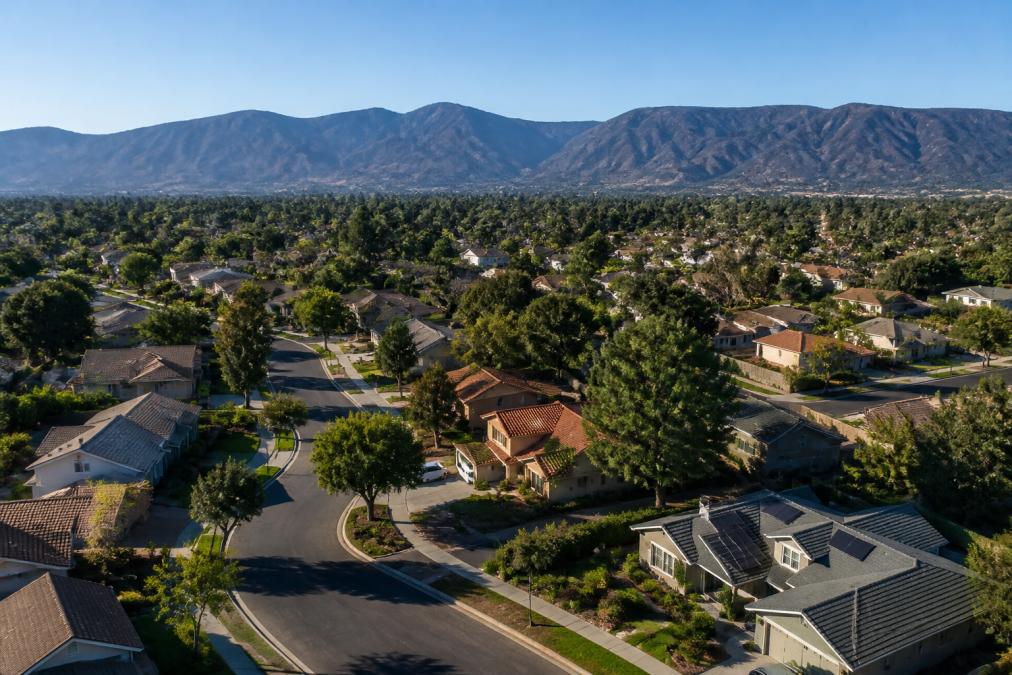 Aerial view of a well-maintained suburban neighborhood in Southern California with tree-lined streets, single-family homes, and mountain ridgelines visible in the background under clear blue skies