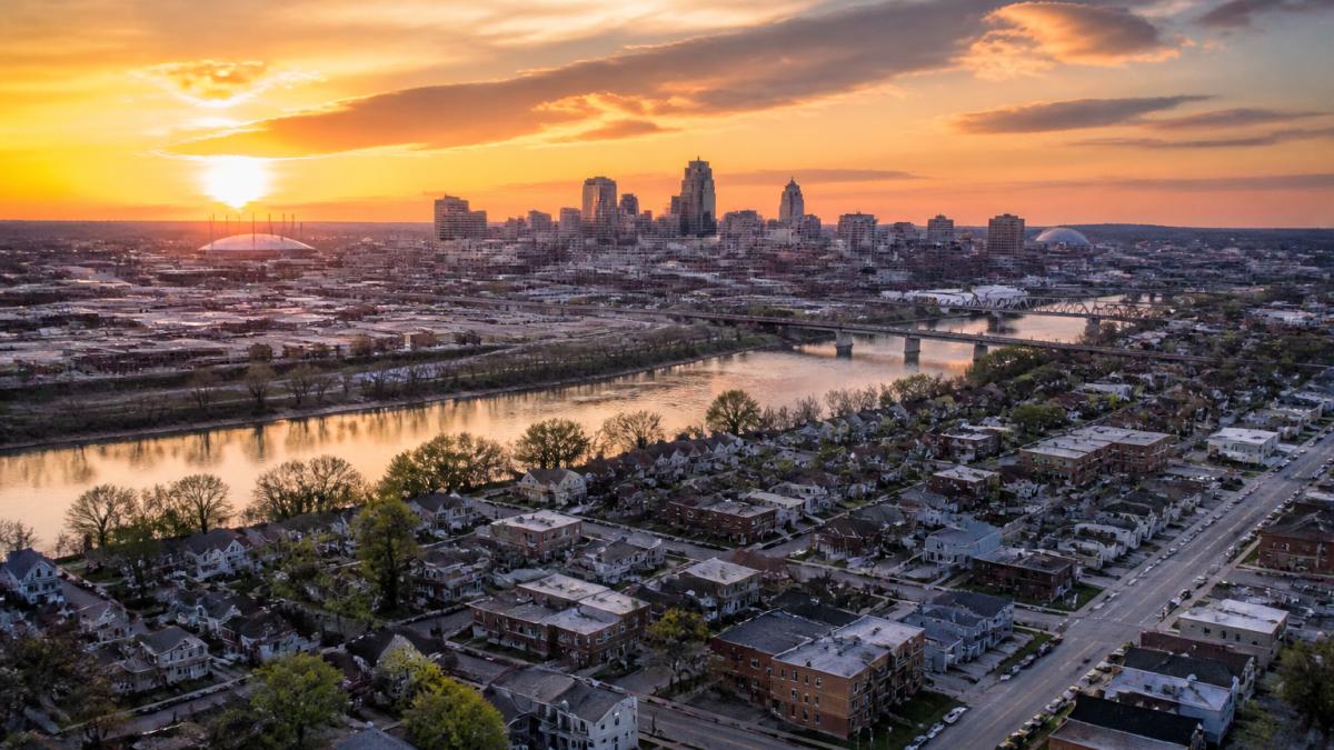 Aerial view of Kansas City skyline at sunset with the Missouri River, showing downtown buildings and neighborhood streets below with warm golden light