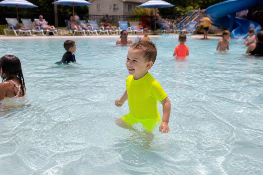 Toddler in bright neon swim set playing in shallow end of crowded community pool, easily visible among other swimmers and pool furniture
