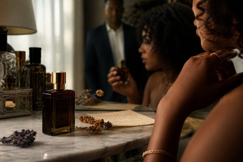 Close-up of a dark amber glass perfume bottle with gold cap on a marble vanity, soft natural window light highlighting the liquid inside, a few dried botanicals and a handwritten note beside it
